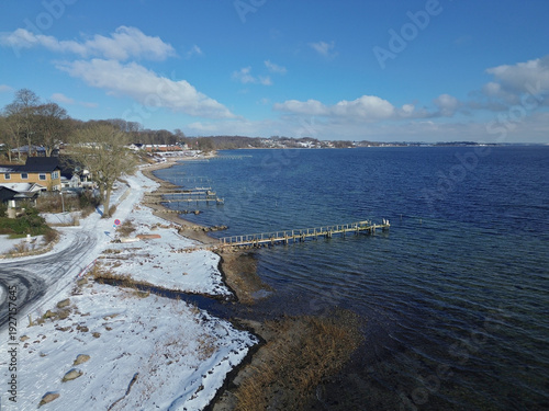 Snowy shoreline, clear shallow water and coastal houses surrounded by trees. The calm fjord, bright sky and visible underwater formations create a serene Nordic winter atmosphere