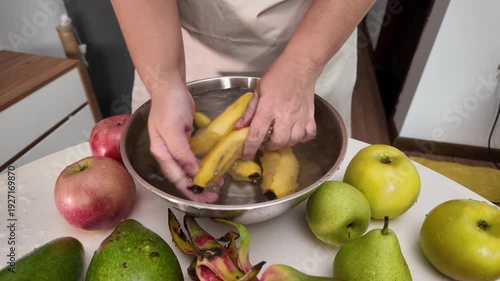 The hands of a woman in an apron standing in front of a table with fruit and washing bananas