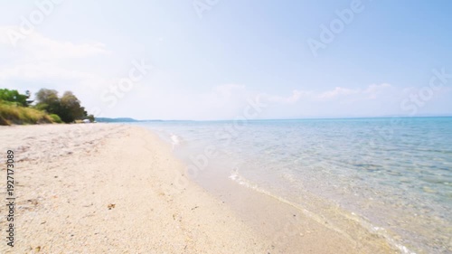A serene seascape showing a clear, calm shoreline with transparent blue water and smooth pebbles visible beneath the surface. The sky is bright and clear with a few clouds. 4K.