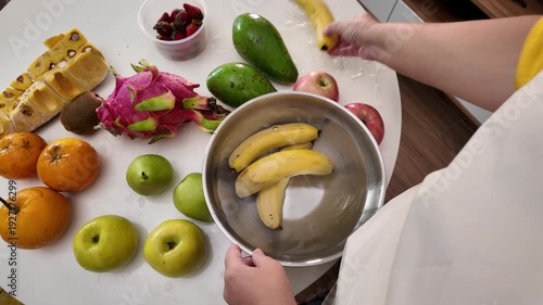 Hands of a woman in an apron standing in front of a table with fruit and washing bananas