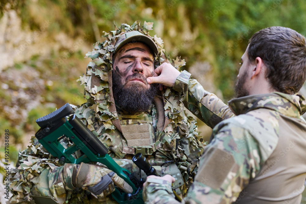 Obraz premium Bearded Soldier In Camouflage Receiving Face Paint Before Field Operation In Forest Training