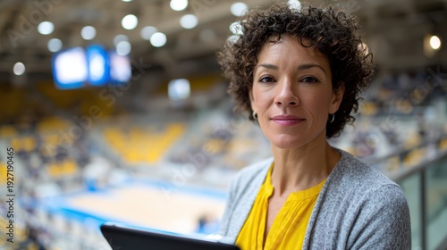 latinx sports executive, a confident latina woman, a top volleyball federation executive, stands focused on a mezzanine, tablet in hand, overseeing a brightly lit arena