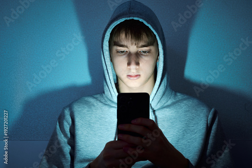 Teenage boy in a hoodie sitting in the dark, illuminated by the glow of his smartphone while looking at the screen