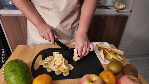 The process of cutting a banana into slices. A woman's hands cut a banana on a chopping board on the kitchen table
