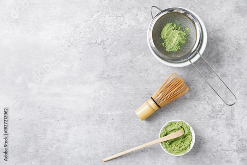 Fotografie Green matcha powder in bowl, scoop, bamboo whisk and sifter on gray textured table, flat lay