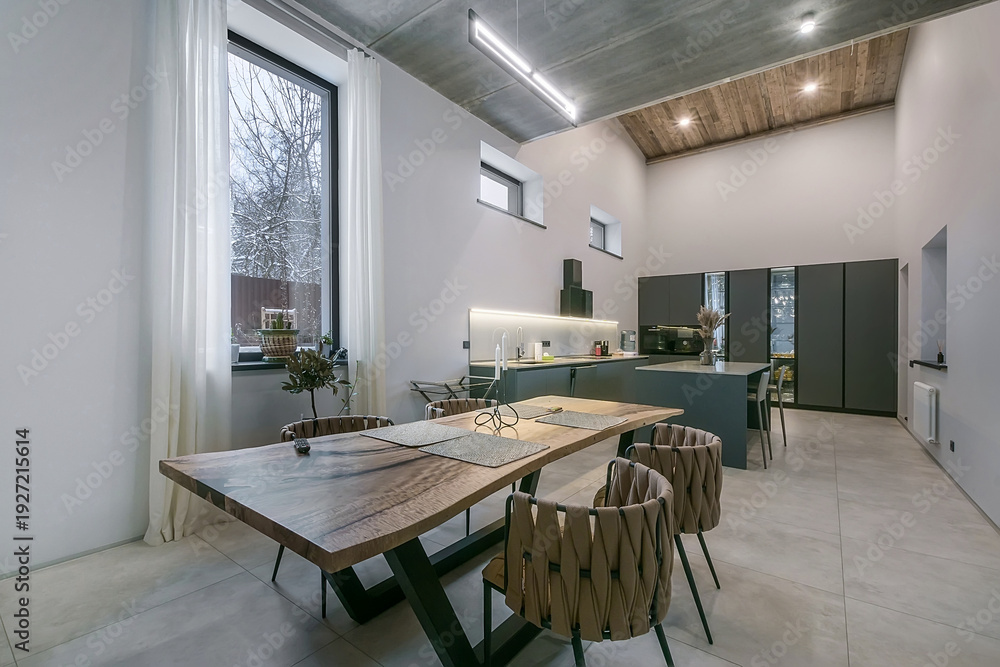 Fototapeta premium A dining table with chairs sits in a modern kitchen. Large windows let in light and show a view of trees outside. The space is open and clean with simple design features