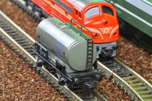 Close-up of a model train tanker car on tracks with a red locomotive behind it. A detailed view of a miniature tanker wagon positioned on a model railway track, with a vibrant red engine visible in