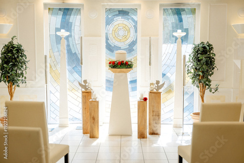 Solemn Funeral Hall with a Memorial Urn on a Pedestal, Surrounded by Candles and Floral Tributes in Soft Natural Daylight