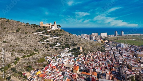 Aerial drone view of Cullera in Spain featuring a hilltop castle and church overlooking the Mediterranean Sea. Detailed coastal landscape with fortress walls, zigzag path and seaside urban skyline.