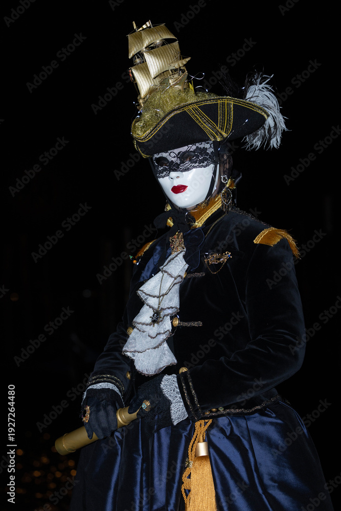 Obraz premium Venice, Italy - February 11, 2026: Elegant masked figure in elaborate costume with gold accents and feathered hat at Venice Carnival night celebration