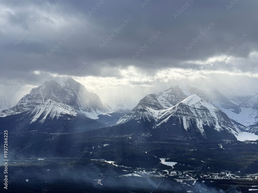 Obraz premium Sun shining through dark clouds above beautiful mountains covered with snow on winter day