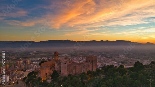 Cinematic aerial view of Cullera in Spain at sunset with castle and church silhouetted against golden light. Mediterranean coastline and mountain backdrop create a dramatic coastal cityscape.