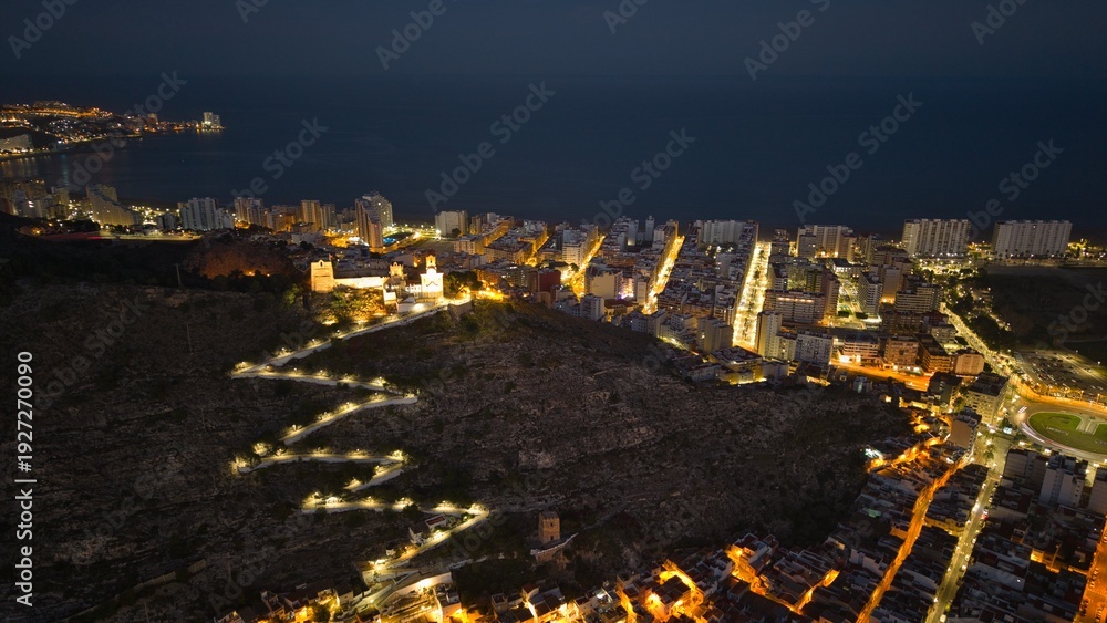 Obraz premium Aerial night view of Cullera in Spain featuring illuminated hilltop castle and glowing city streets along the Mediterranean coast. Detailed coastal skyline with fortress walls and seaside urban landsc