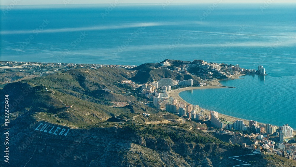 Obraz premium Wide aerial panorama of Cullera in Spain showing the Mediterranean coastline, river mouth and mountain ridge. Detailed drone view of seaside city, beaches and surrounding landscape.