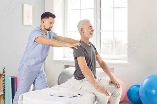 Young doctor osteopath or physiotherapist helping senior mature male patient sitting on the couch in modern rehab clinic with healing back pain. Physiotherapy and physical rehabilitation concept.