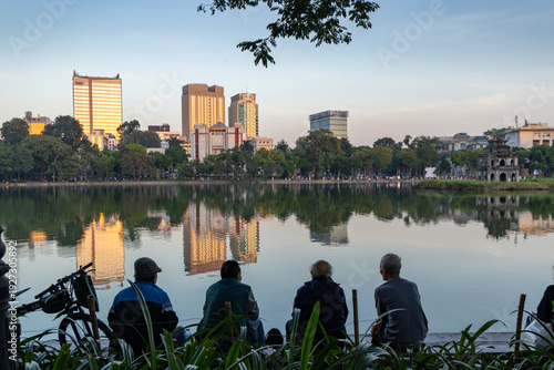 Four men sit and relax by Hoan Kiem Lake in Hanoi, Vietnam, enjoying the sunset and the reflections of the city skyline on the water. They are taking in the view.