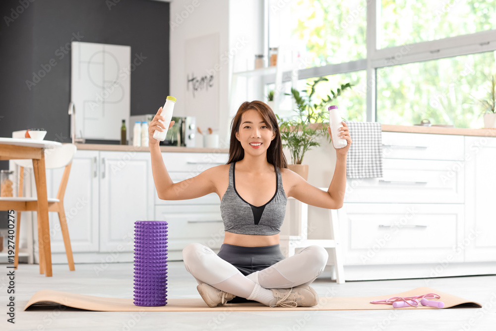 Fototapeta premium Sporty young Asian woman with bottles of tasty yogurt sitting on yoga mat at home