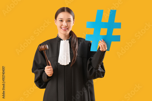 Young female judge in robe with gavel and hashtag sign on yellow background
