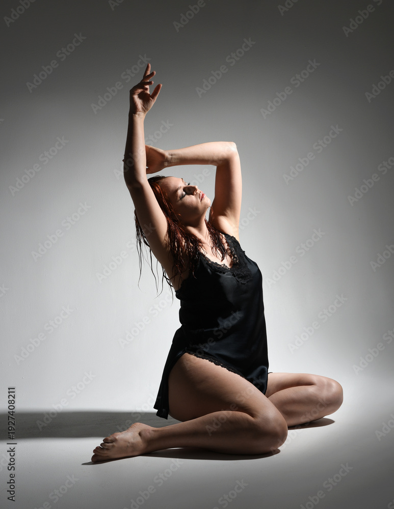 Obraz premium full length portrait of beautiful girl model, wet messy hair, simple black silk robe gown, bruised leg. sitting pose with emotive hand gesture reaching. isolated figure studio background dark shadows