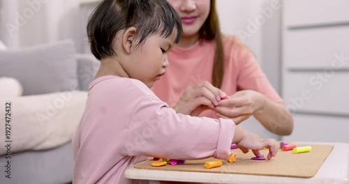 Asian preschool daughter concentrating on shaping clay while mother provides guidance during home learning activity both seated at low table filled with colorful materials encouraging independent crea