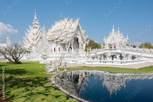The White Temple, Wat Rong Khun, in Chiang Rai, northern Thailand. Iconic contemporary Buddhist temple with intricate white architecture and reflective pond under blue sky. Famous Thai landmark, relig
