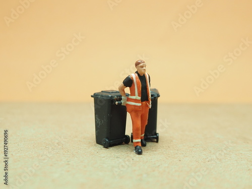 Mini toy of action figure with blurred background. Miniature diorama photography concept design. Minifigure of cleaning crew character wearing orange suit with truck and garbage bin.