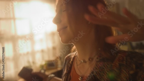 Candid close up shot of young woman enjoying music and dancing at home using wireless earbuds holding phone in hand with warm cinematic lighting, copy space