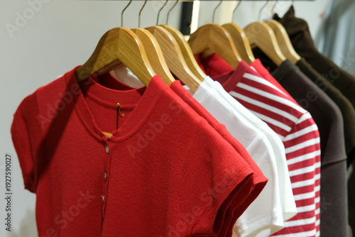 Row of stylish red and white shirts hanging on wooden hangers in a modern clothing store. Striped and chocolate brown T-shirts displayed on rack with soft lighting. Casual wear collection