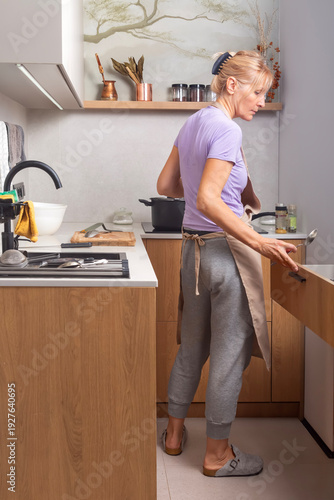 A woman in an apron, cooking soup, takes a spoon out of a drawer and closes it, in a small stylish kitchen. Competent organization of a small workspace in the kitchen