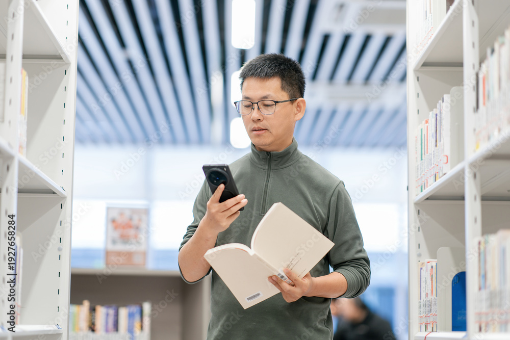 Fototapeta premium Young man in a library holding an open book while checking his smartphone