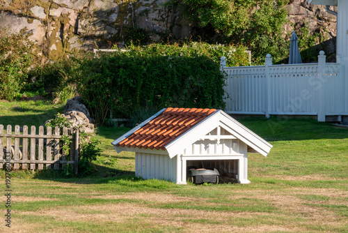 Robotic lawn mower parked inside small wooden garden shelter on residential lawn in Hamburgö, Bohuslän, Sweden
