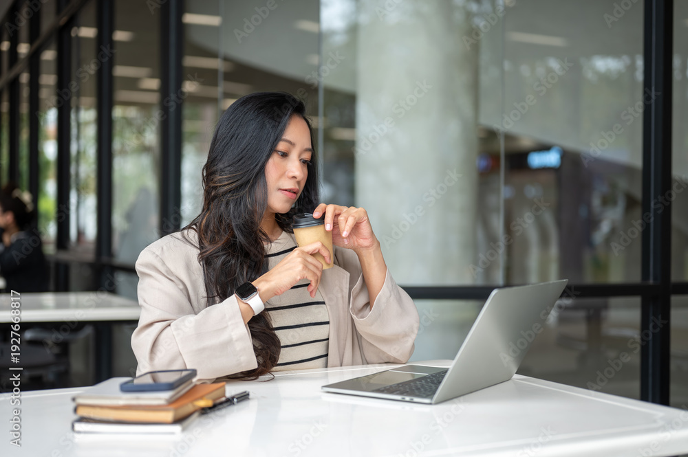 Fototapeta premium Asian woman holding coffee looking at laptop aside book and phone sitting at table in cafe building.