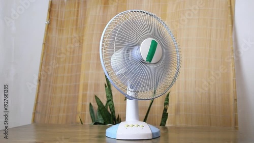 Electrical stand fan oscillating on a wooden surface for cooling and ventilation, with a natural bamboo curtain in background