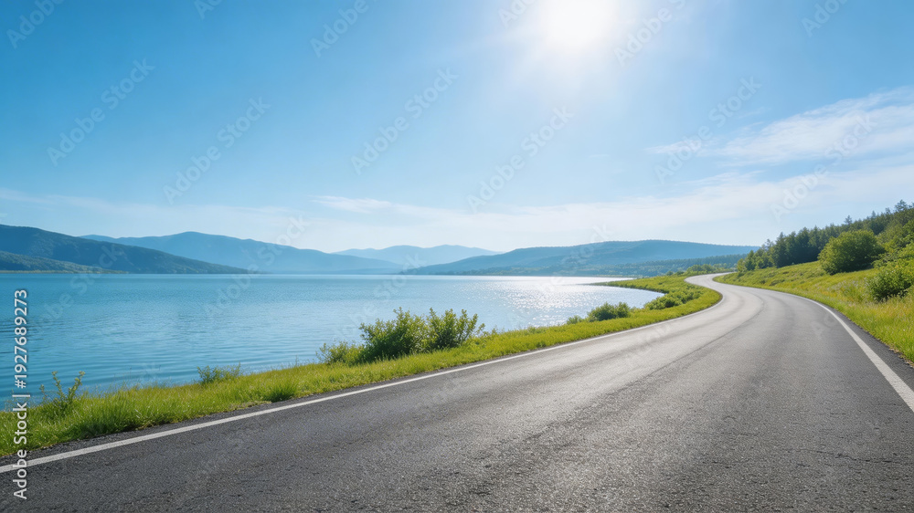 Naklejka premium A smooth, curved road runs alongside a calm lake, flanked by lush greenery. Distant mountains rise under a bright, clear sky, creating a serene, picturesque landscape ideal for travel and adventure.