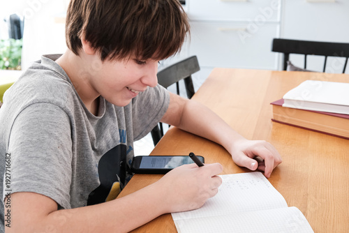 A teenager does his math homework, writes down examples in a notebook, and uses his smartphone for educational purposes. copying ready-made homework solutions.  digital learning.