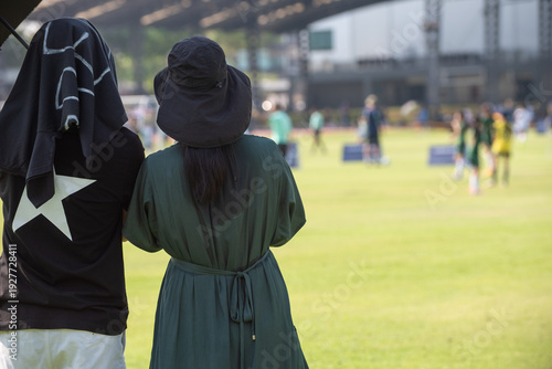 Candid back view of a supportive Asian mothers cheering for their son during a youth soccer match at school.