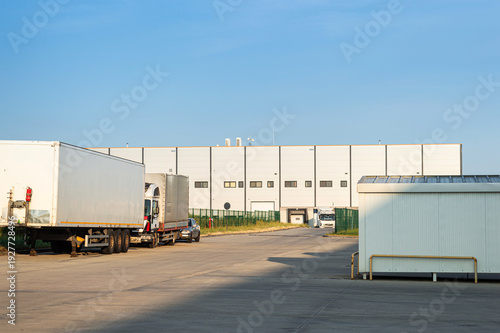 Fotografi Industrial warehouse exterior with loading docks and parked semi-trailer trucks under clear blue sky