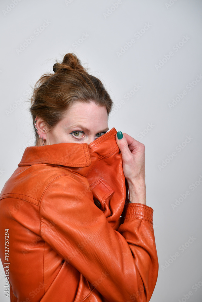 Fototapeta premium Frauenportrait im Studio mit orangefarbener Lederjacke. Das Model zieht den Kragen hoch und blickt intensiv in die Kamera – modernes Fashion-Statement mit selbstbewusster Attitude.