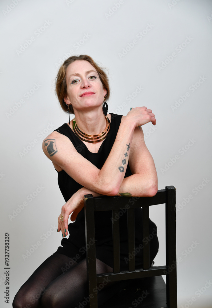 Naklejka premium Selbstbewusstes Frauenportrait im Studio. Model in schwarzem Outfit mit Tattoos, sitzend auf einem Stuhl – minimalistisches Setting mit klarer Haltung und moderner Ästhetik.