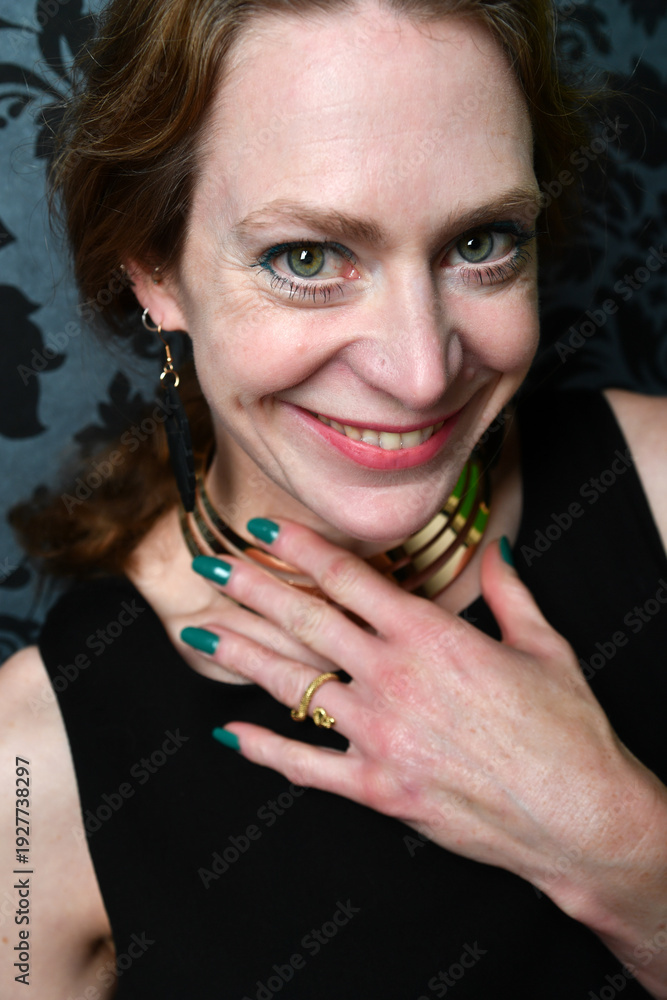 Obraz premium Ausdrucksstarkes Frauenportrait mit direktem Blick und lebendigem Lächeln. Schmuckdetails und grün lackierte Nägel setzen Akzente – selbstbewusstes, modernes Studiofoto.