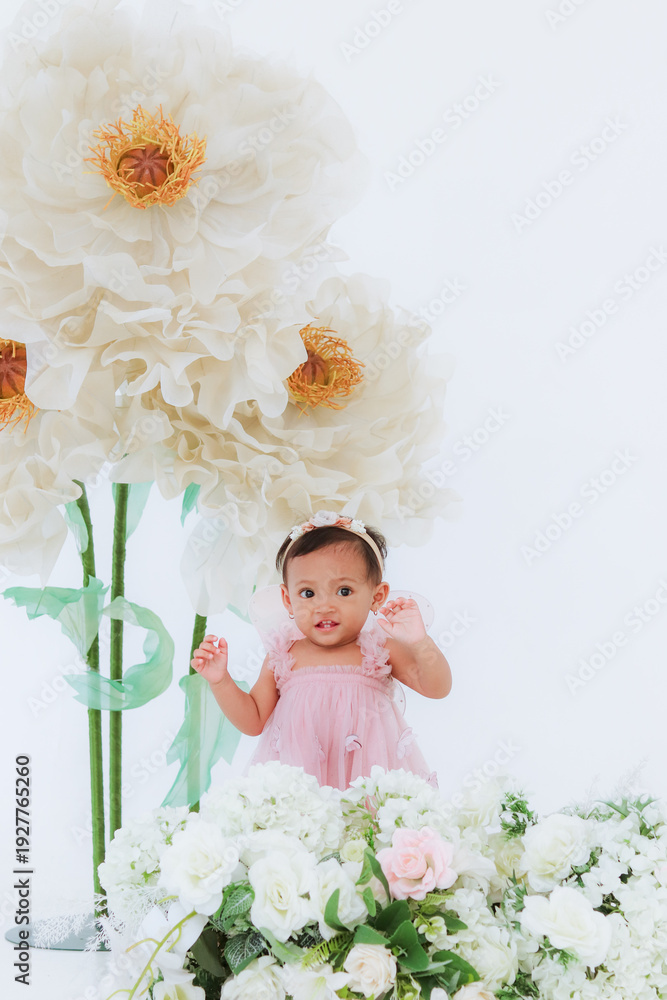 Fototapeta Adorable baby girl in pink dress standing near giant white paper flowers, soft pastel studio portrait with elegant floral backdrop and copy space.