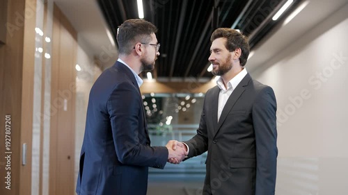 Two confident businessmen in formal suits greet each other with a handshake in a corridor of modern business center. Colleagues engaged in teamwork, communicate, discuss work issues, exchanging ideas