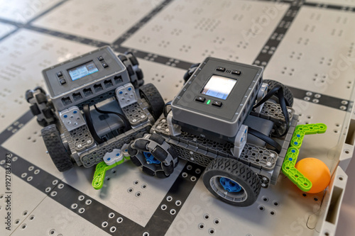 Two small robots are positioned on a grid-patterned floor, ready for a task involving a ball.
