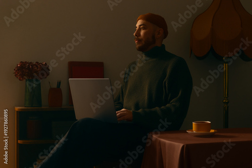 Fotografie Focused man in a beanie and turtleneck working on a laptop in a cozy, dimly lit vintage interior with warm lighting