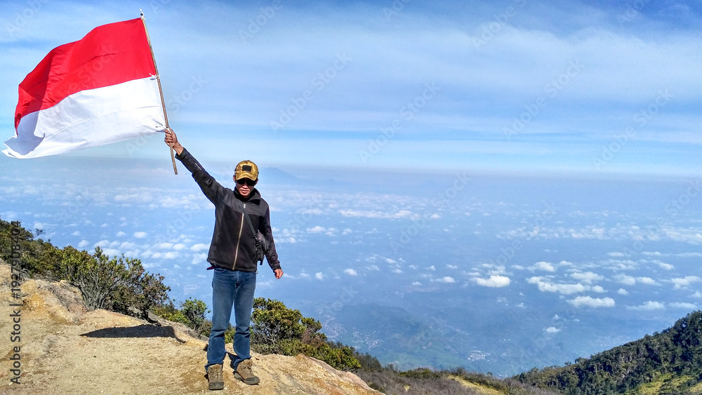 Obraz premium Hiker proudly raises Indonesian flag on mountain peak above clouds