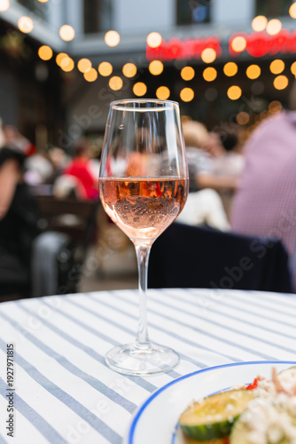 Close-up of a glass of rose wine on a striped tablecloth at an outdoor cafe terrace in the evening with blurred people and bokeh lights in the background.
