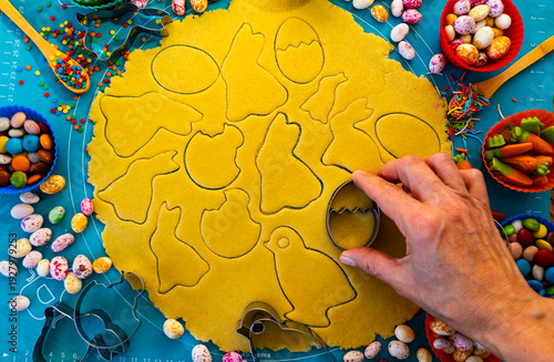 A hand carefully uses cookie cutters to cut out festive Easter cookies in the shapes of bunnies, eggs, and chicks from rolled‑out shortcrust dough.