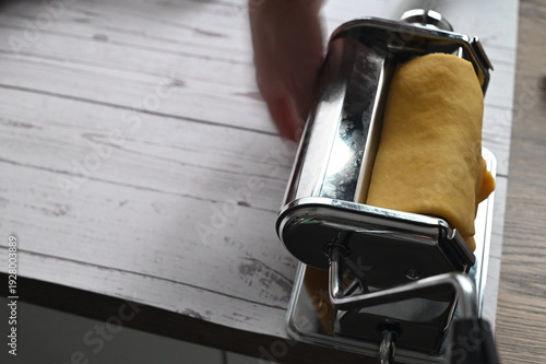 Pasta machine rolling homemade dough on a white table in a home kitchen.