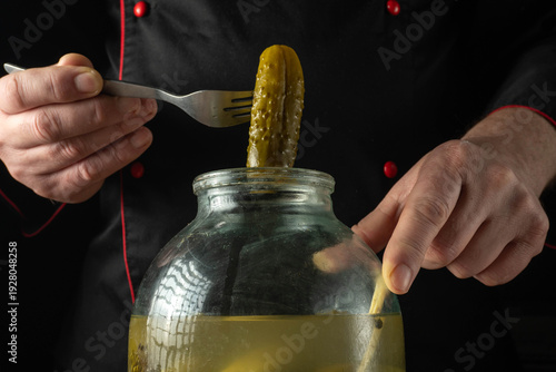 A chef uses a fork to lift a cucumber pickle from a large glass jar filled with brine. The setting is a kitchen with a dark background. The jar contains several pickles