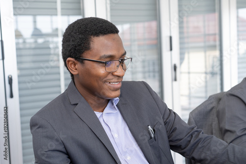 A black man in a suit sits smiling at work.
